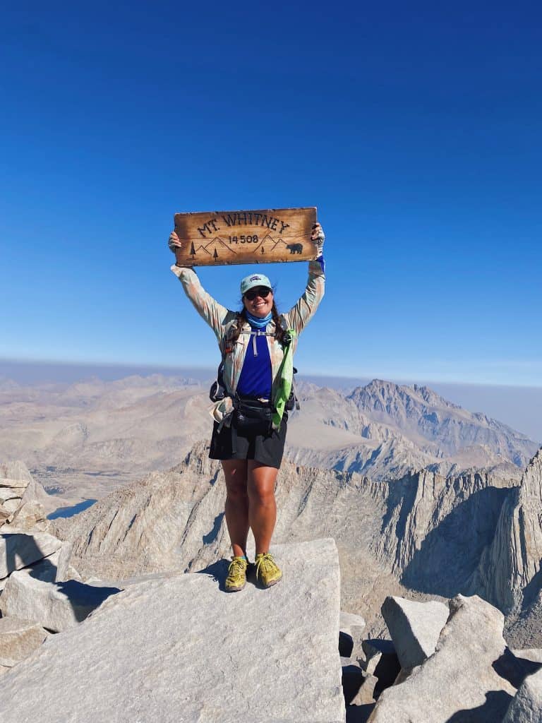 author Emily standing on the summit of mt Whitney holding a sign reading mt Whitney 14,505 feet