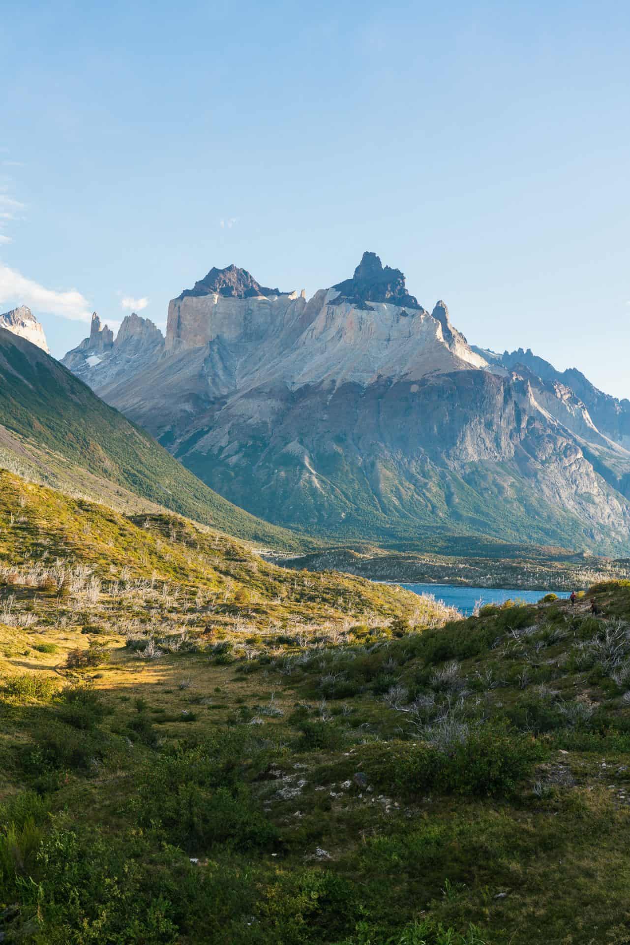 Cuerno Principal in Torres del Paine National Park as viewed from the west. Sloping green hillsides with shrubs and small burned trees dot the foreground as the mountain massif lies large in the background.