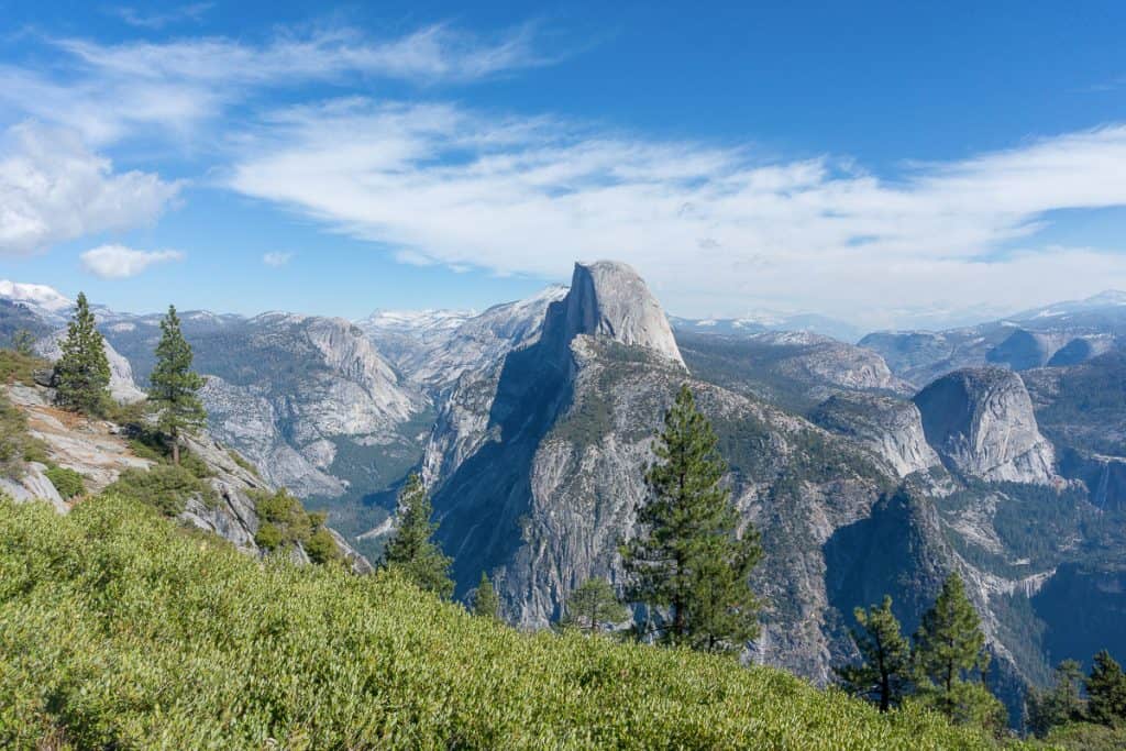 half dome sitting upon large granite slope with smaller granite features in the background and trees and shrub in foreground. Partly cloudy blue skies above