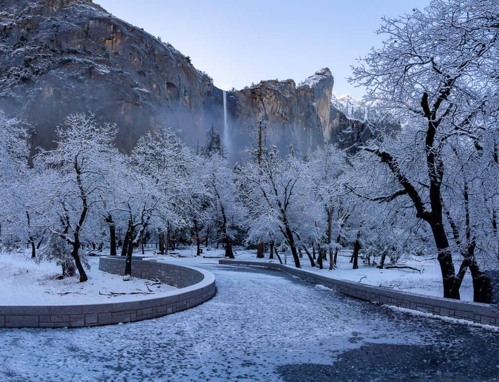 Snowy stone path with leafless snowy trees and a waterfall in the background flowing over a granite wall in Yosemite national park in winter