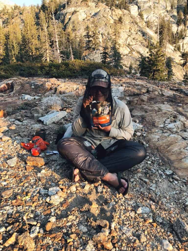author Emily sitting on the ground eating a dehydrated meal while wearing a bug head net in northern yosemite