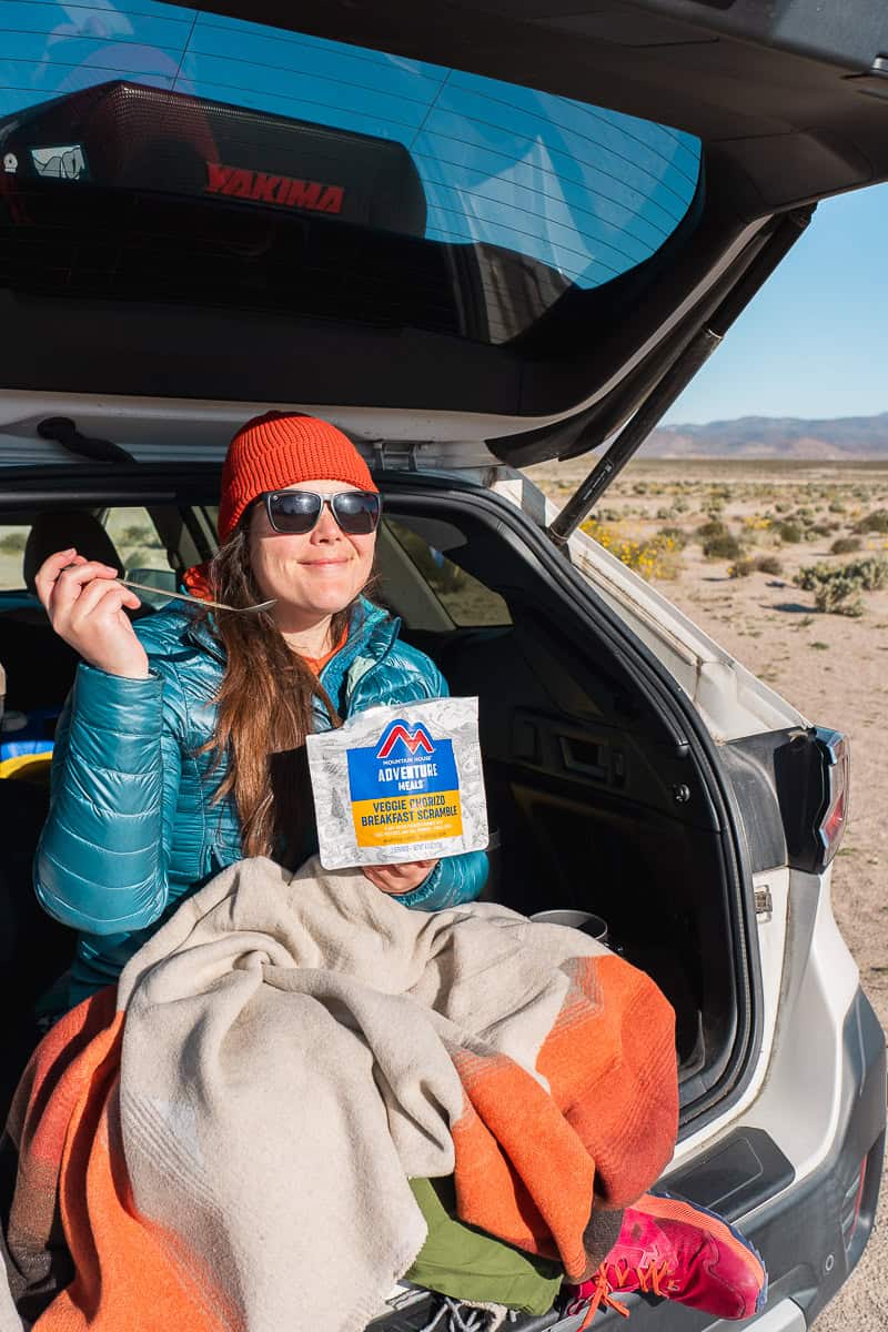 A woman sitting in the trunk of a car with a blanket and a freeze dried bag meal