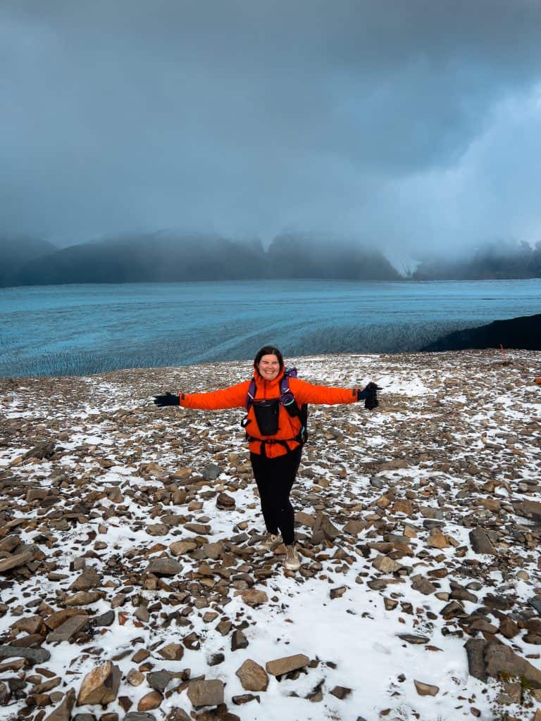 author Emily in red rain jacket standing with arms outstretch at the top of John Gardner pass with glacier grey in background