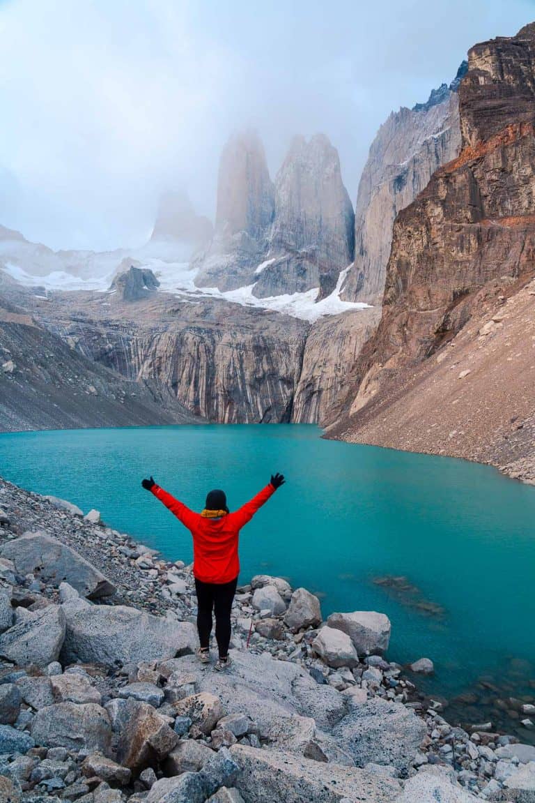 woman hiker in red standing with arms raised looking at mountains and a blue lake