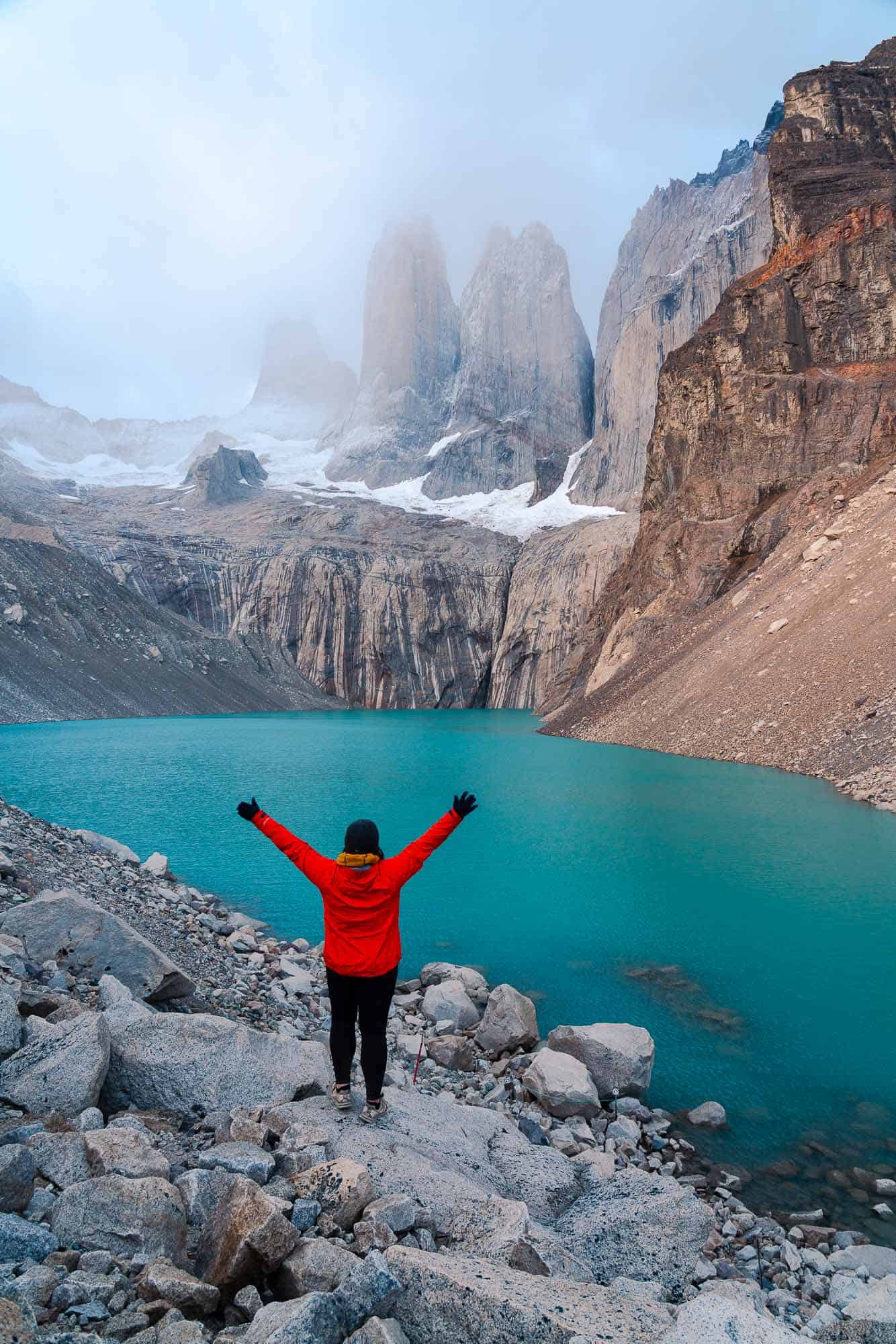 woman hiker in red standing with arms raised looking at mountains and a blue lake