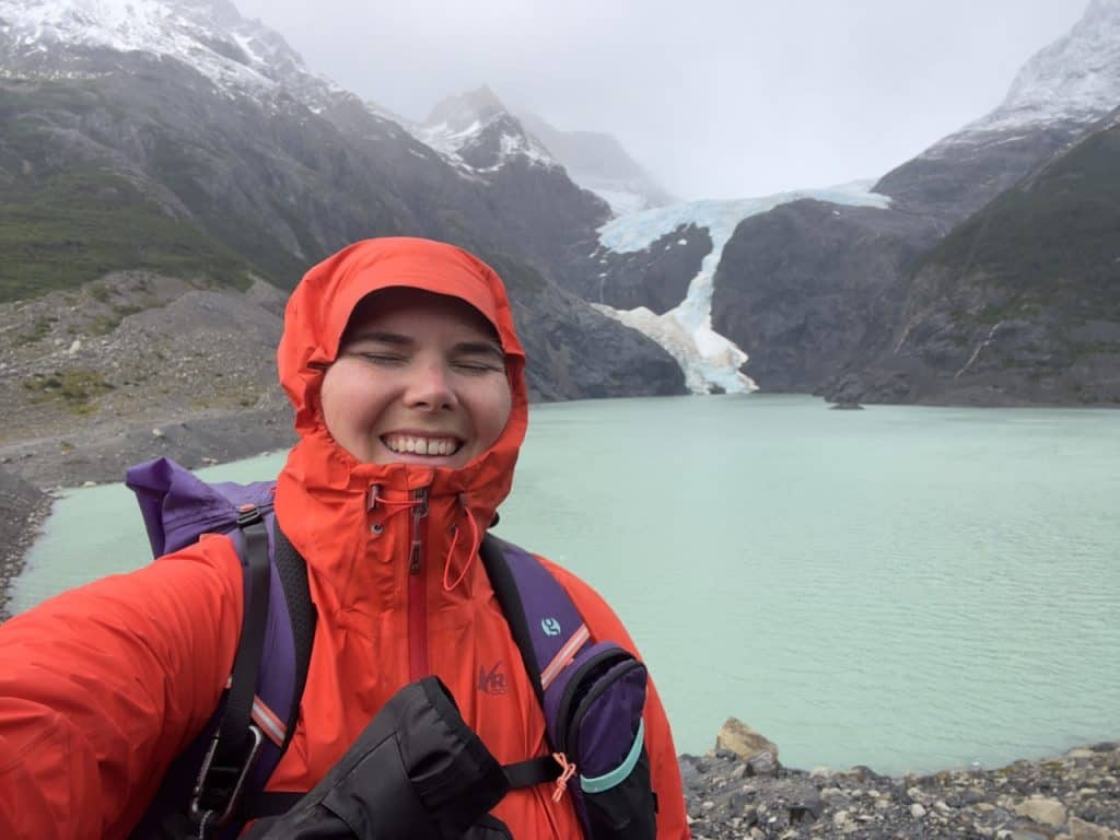 Emily smiling with eyes closed wearing rain jacket with hood on and backpack in front of a blue glacial lake