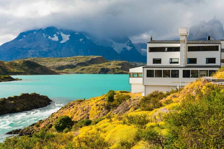 Serene mountain lake retreat with modern house, lush yellow foliage, and dramatic cloudy mountain backdrop.