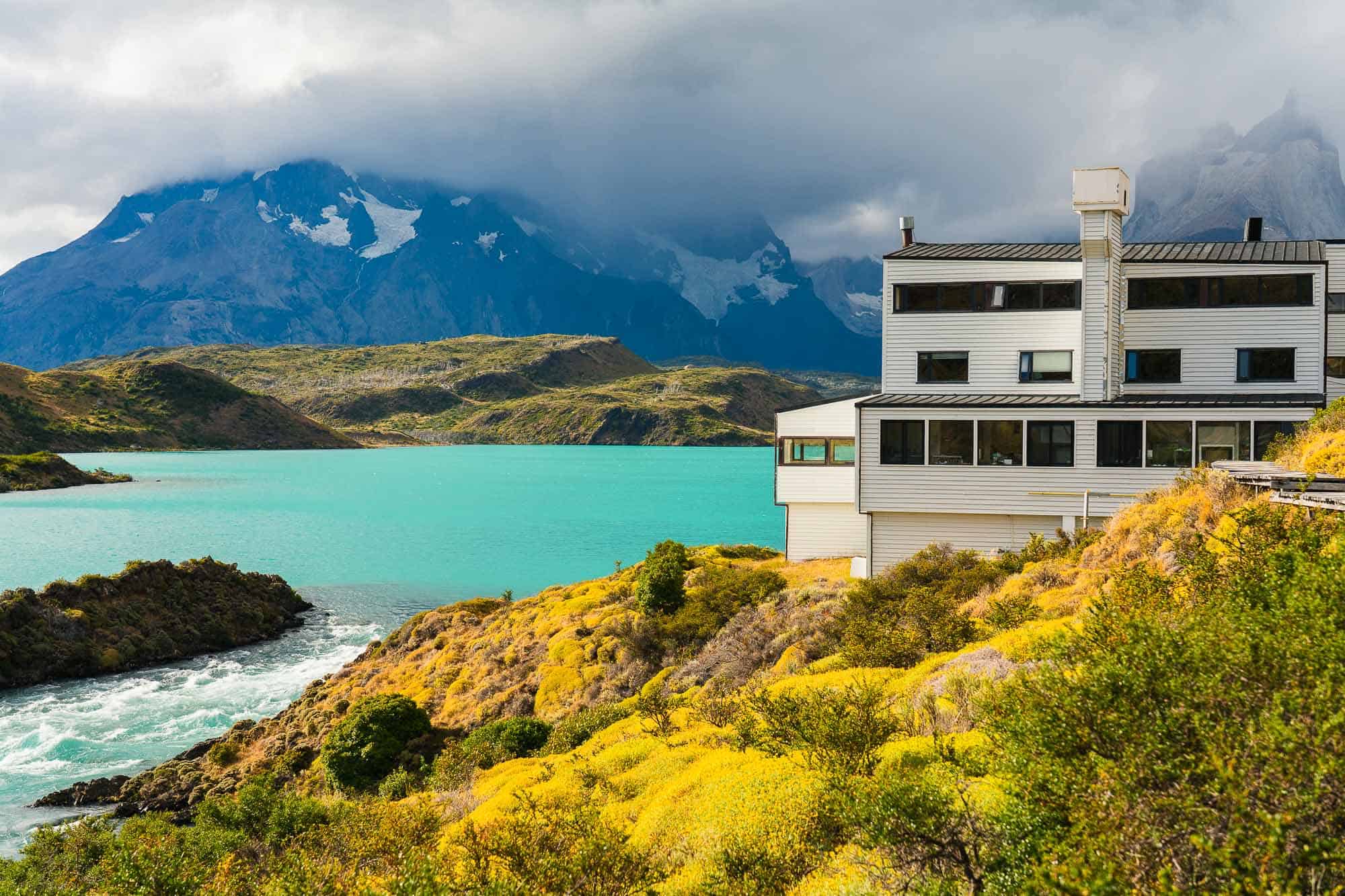 Serene mountain lake retreat with modern house, lush yellow foliage, and dramatic cloudy mountain backdrop.