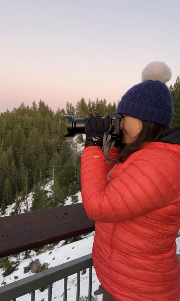 author in pink puffy jacket wearing gloves while using camera in the snow