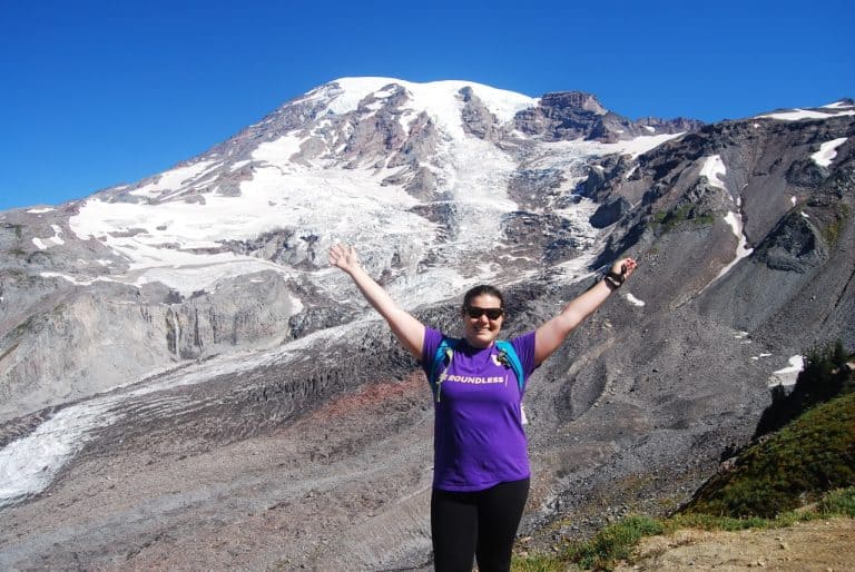 author Emily standing in front of mt rainier with her arms raised up wearing a cotton UW t shirt