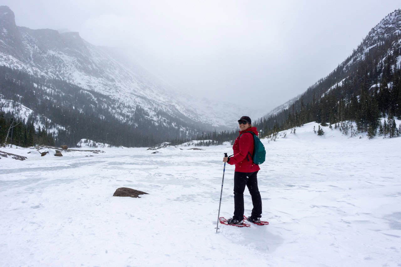 A woman snowshoeing in a scenic winter mountain landscape with snowy peaks and forests.