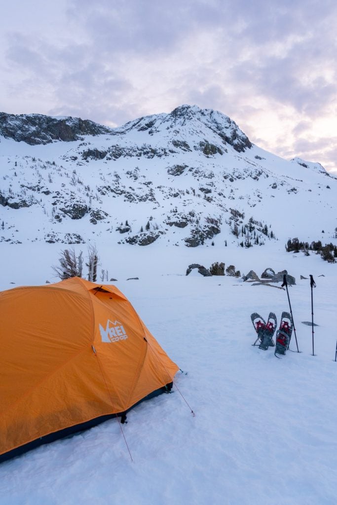 orange tent pitched on snow with snowshoes and trekking poles placed next to it and a snowy mountain looms behind
