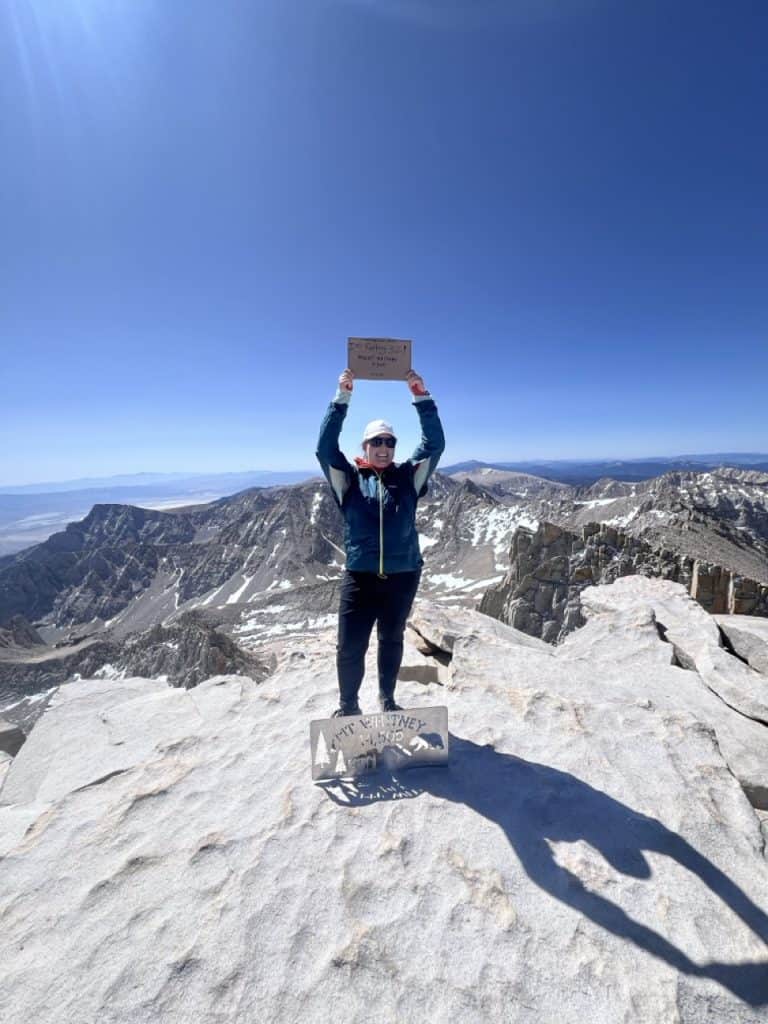 Person standing on Mount Whitney summit peak holding a sign.